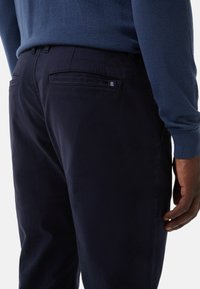 Navy trousers with a smooth texture, featuring two back pockets and a small logo on the right pocket. Worn with a blue sweater.