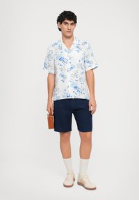 Camicia bianca a maniche corte con motivo floreale blu, pantaloni corti blu navy, scarpe da ginnastica beige e una piccola borsa marrone. Sfondo semplice.