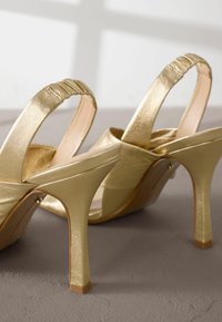 Gouden slingback hakken met een glanzende metallic afwerking, voorzien van een hoge stiletto hak, gestructureerde banden en een ronde neus.