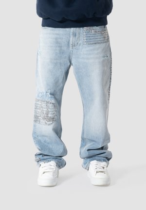 Lyseblå slidte jeans med dekorative lapper på lår og lomme, brugt med hvide sneakers og en mørk top.