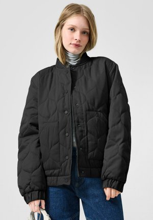 Jonge vrouw die een zwarte gewatteerde jas draagt over een lichtgrijze coltrui, blauwe spijkerbroek, en een kleine zilveren handtas vasthoudt tegen een effen grijze achtergrond.