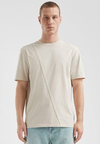 Beżowa koszulka z krótkim rękawem z ukośnym szwem, wykonana z miękkiej bawełny. Prosty okrągły dekolt i luźny krój.