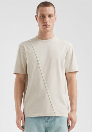 Beige T-Shirt mit kurzen Ärmeln und diagonaler Nahtdetails, aus weichem Baumwollstoff gefertigt. Schlichter Rundhalsausschnitt und lockere Passform.