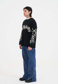 Czarny sweter dzianinowy z białym napisem oraz wzorami w spirale na rękawach, połączony z luźnymi niebieskimi spodniami w paski i czarnymi butami.