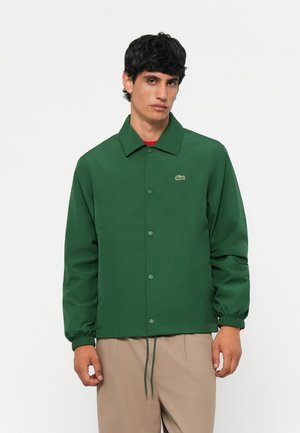 Green button-up jacket with a collar, elastic cuffs, drawstring hem, and small logo detail. Smooth texture, casual fit.