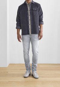 Chemise en velours côtelé gris foncé à boutons, associée à une chemise à carreaux à boutons et un jean gris clair. Des baskets grises complètent la tenue.