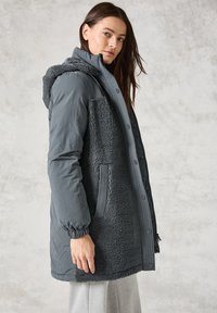 Manteau long gris avec une partie supérieure lisse et résistante à l'eau et un côté en texture polaire, doté d'une capuche, de poignets élastiques et de poches latérales.