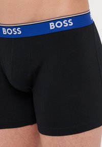 Crne pamučne bokserske gaće s plavim elastičnim pojasom. Pojas sadrži bijeli tekst "BOSS" i suptilan prugasti detalj.