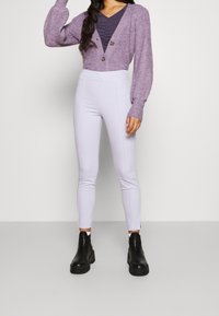 Lila stickad cardigan med två knappar, bärs över en mörk topp, i kombination med ljuslila tighta byxor och svarta ankelskor.