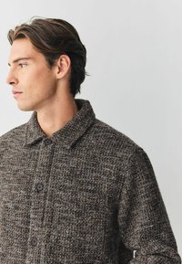 Chaqueta texturada marrón con cuello de solapa, que presenta un cierre de botón y un sutil patrón tejido. Tejido suave con un ligero brillo.