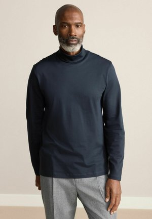 Navy turtleneck long-sleeve shirt made of soft fabric, featuring a fitted design and minimal detailing, paired with gray trousers.