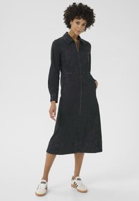 Robe chemise en denim foncé avec des manches longues, une fermeture éclair à l'avant, des poches poitrine et une longueur mi-jambe. Portée avec des baskets blanches et noires.