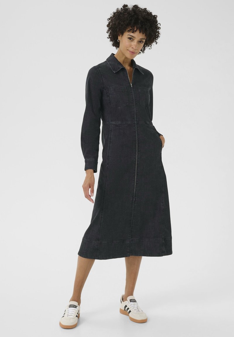 Robe chemise en denim foncé avec des manches longues, une fermeture éclair à l'avant, des poches poitrine et une longueur mi-jambe. Portée avec des baskets blanches et noires.