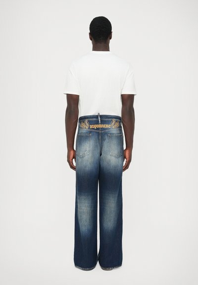 Uomo che indossa jeans larghi sbiaditi blu con logo ricamato e design sulla vita, abbinati a una t-shirt bianca a maniche corte.