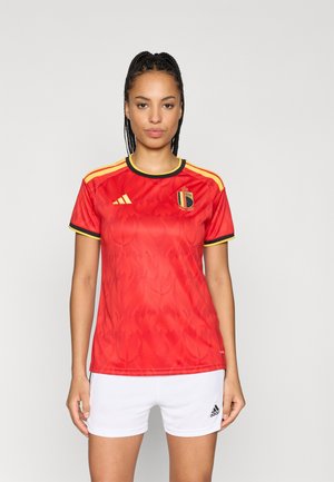 Maillot de football rouge avec un motif de flammes, accents noirs et jaunes, logo Adidas, accompagné d'un short blanc, avec un logo sur le short.