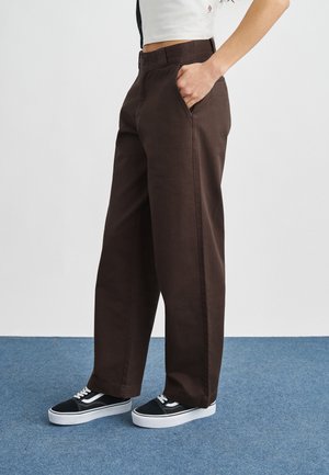 Bruine wijde broek gemaakt van stevig stof, met zijzakken en een gladde afwerking, gecombineerd met zwarte sneakers met witte accenten.