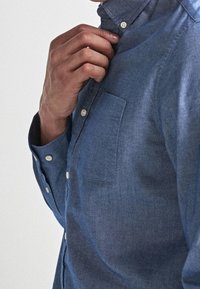 Man die de kraag van een blauwe chambray overhemd met lange mouwen, witte knopen en een borstzak dichtknopt.