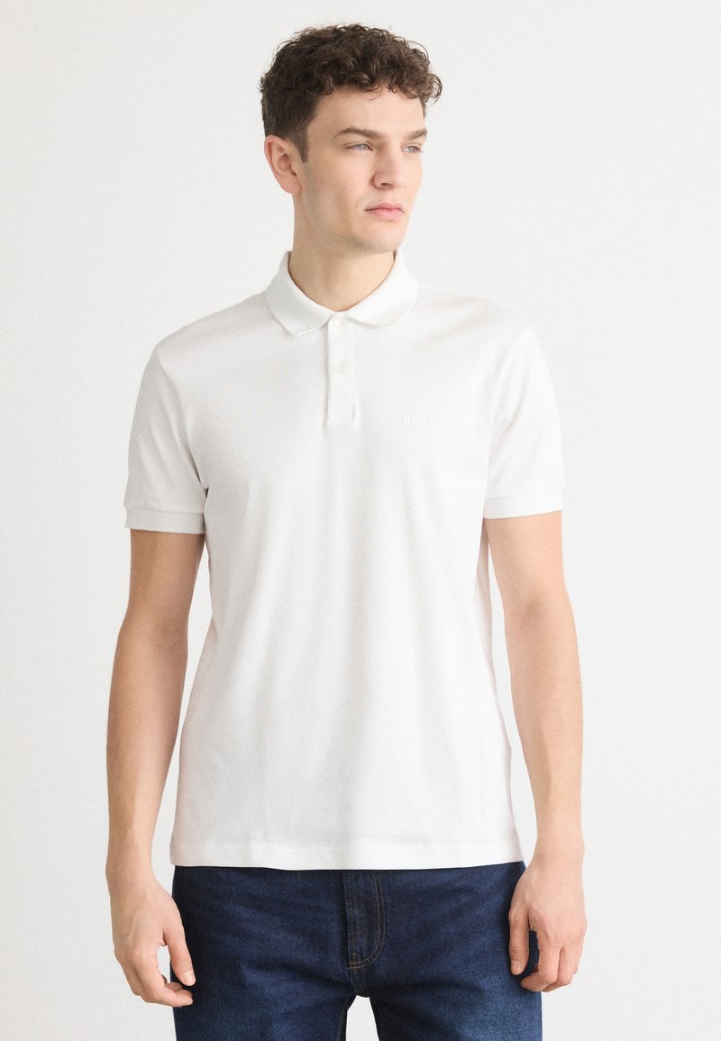 Vit polo-shirt i lätt tyg, med klassisk krage, tre knappar och korta ärmar. Diskret logotypdetalj på bröstet.