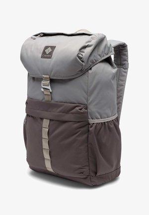 Jeune femme en chemise noire et short vert portant un grand sac à dos de randonnée bicolore gris, debout de profil sur fond blanc.