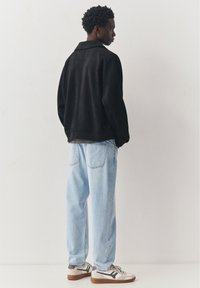 Jonge man opzij kijkend draagt een zwart jack, lichtblauwe spijkerbroek en witte sneakers tegen een effen witte achtergrond.