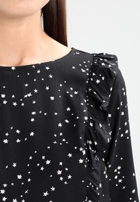 Haut en tissu noir avec un motif d'étoiles blanches, présentant une accentuation froncée le long de l'épaule et un col arrondi. Texture lisse.