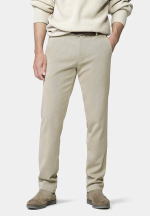Mann trägt beige Hose, hellbeigen Pullover mit hochgekrempelten Ärmeln, braunen Gürtel und braune Wildlederschuhe, steht mit einer Hand in der Tasche.