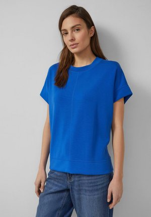 Jonge vrouw met stijlbruin haar, gekleed in een felblauwe top met korte mouwen en een blauwe spijkerbroek, tegen een effen grijze achtergrond.