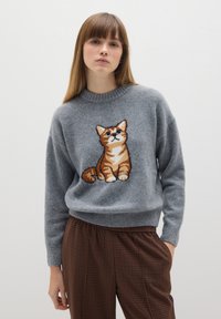 Grijze gebreide trui met een geborduurd ontwerp van een oranje tabby kat op de voorkant. De stof heeft een zachte textuur en wordt gecombineerd met bruine geruite broeken.