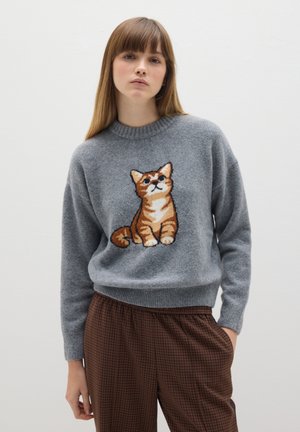 Maglione grigio a maglia con un design ricamato di un gatto arancione tigrato sul davanti. Il tessuto ha una consistenza morbida, abbinato a pantaloni a quadri marroni.