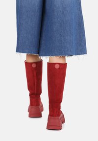 Stivali alti fino al ginocchio in suede rosso con una superficie texturizzata, accenti rotondi con logo nella parte superiore e suole in gomma spesse in tonalità abbinata.