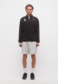 Νεαρός άνδρας που στέκεται κρατώντας μικρούς μαύρους αλτήρες, φορώντας μαύρο μπουφάν με φερμουάρ, γκρι σορτς, λευκές κάλτσες και μαύρα αθλητικά παπούτσια σε λευκό φόντο.