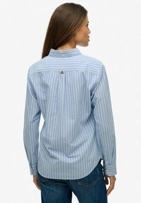 Niebiesko-biała koszula w paski z długim rękawem, kołnierzykiem, zapinana na guziki z tyłu i z małym logo blisko kołnierzyka. Prezentuje luźny krój.