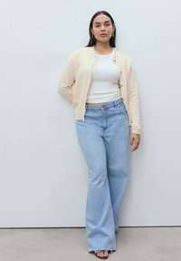 Cardigan texturizado en color crema con cierres de botón, combinado con una camiseta blanca ajustada y jeans acampanados en azul claro. Sandalias de tacón negras completan el look.