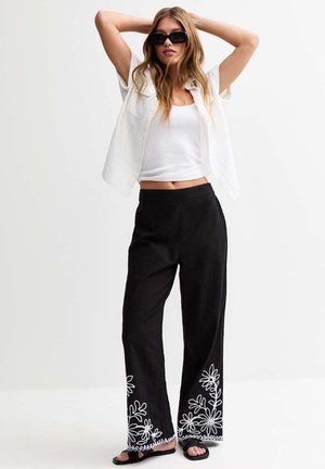 Black wide-leg pants with white floral embroidery at the hems, paired with a white cropped top and open, lightweight white jacket.