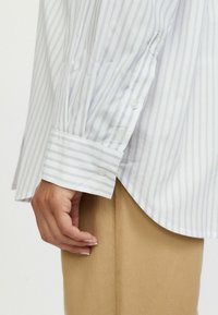 Cămașă albă cu dungi verticale de un mov deschis, cu manșetă cu nasturi și detaliu brodat. Croială lejeră cu tiv curbat.