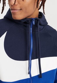 Námořnicky modrá a bílá mikina na zip s vysokým límcem, se vzorem rozdílných barevných bloků a texturovanou látkou, zdůrazňující modrý zip a šňůrky na stažení.