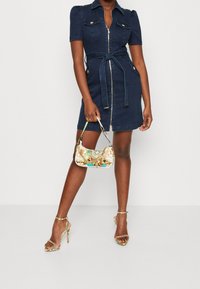 Vestido corto de mezclilla oscura con cierre de cremallera, mangas abullonadas y cintura ceñida con cinturón. Combinado con un bolso de mano de patrón metálico y tacones con tiras.