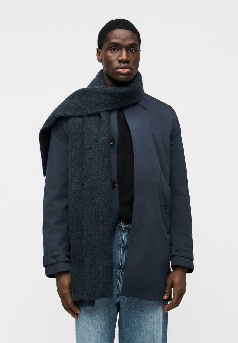 Σκούρο μπλε παλτό από ελαφρύ υλικό, σε συνδυασμό  με παχύ, υφασμάτινο σκούρο κασκόλ και τζιν παντελόνι ανοιχτού πλυσίματος. Απλός, λειτουργικός σχεδιασμός.