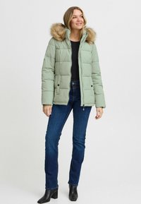 Chaqueta verde acolchada con capucha forrada de piel; presenta un diseño acolchado, bolsillos frontales y cierre con cremallera. Combinada con jeans azules y botas negras.