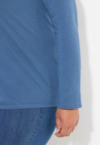 Modrý dlouhý rukáv top vyrobený z měkké látky s uvolněným střihem, s kulatým výstřihem a zakončenými lemy, spárovaný s tmavými džíny.