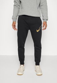 Man wearing black Nike sweatpants with gold and white swoosh logos and "Just do it." text, paired with white sneakers and gray sweatshirt.
