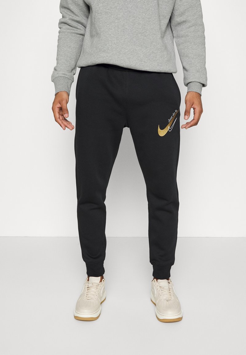 Man wearing black Nike sweatpants with gold and white swoosh logos and "Just do it." text, paired with white sneakers and gray sweatshirt.