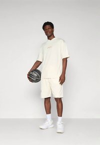Jovem vestido com roupa desportiva bege segura uma bola de basquetebol preta, em pé contra um fundo cinza claro liso.
