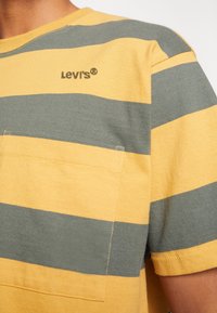 Chemise rayée jaune et grise horizontale avec une poche poitrine et un petit logo Levi's sur le côté supérieur gauche près de l'épaule.