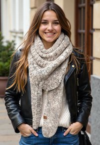 Beżowy, dziany szalik z teksturowanym wzorem, noszony na kremowym swetrze, w połączeniu z czarną skórzaną kurtką, na zewnątrz.