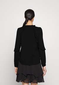 Svart topp med långa ärmar, ruffle-detaljer längs axlarna och halsringningen, i kombination med en lager på lager svart kjol. Slät tygstruktur.