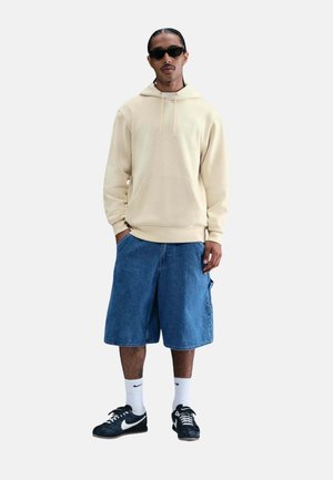 Man draagt een beige hoodie, blauwe spijkershorts, witte Nike-sokken, zwarte Nike-sneakers en zwarte zonnebril, staand met handen in de zakken.