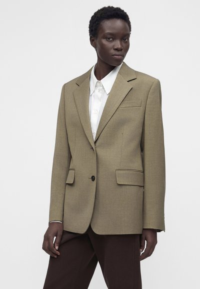 Γυναίκα με μπεζ σακάκι με ένα κουμπί πάνω από λευκό πουκάμισο με γιακά και σκούρο καφέ παντελόνι, στέκεται μπροστά σε απλό φωτεινό φόντο.