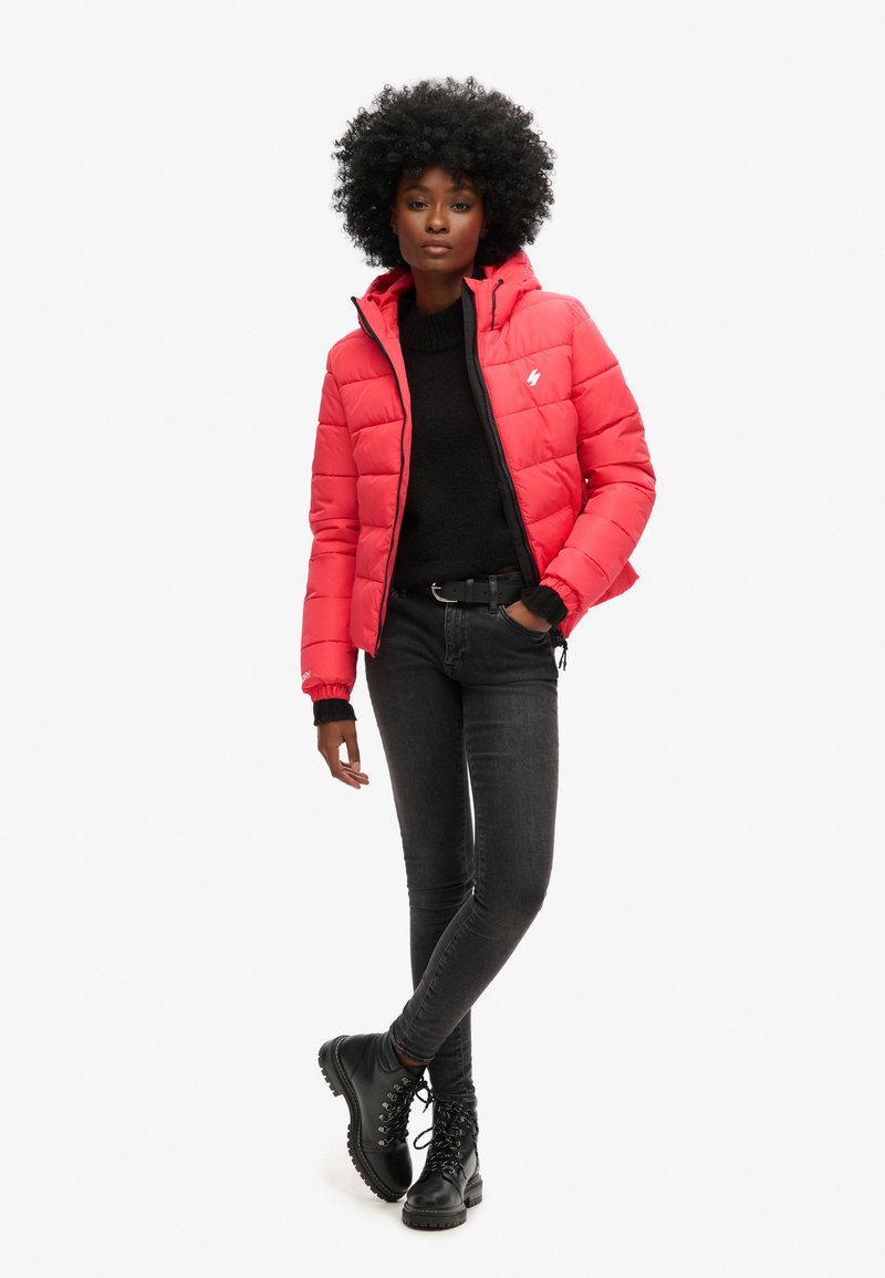 Czerwona kurtka puchowa z kapturem, pikowana, z elastycznymi mankietami. Połączona z czarnym swetrem, dopasowanymi dżinsami i czarnymi butami na sznurówki.