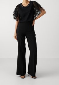 Top negro con mangas transparentes y en capas con lunares, y un escote adornado con cuentas, combinado con pantalones negros de talle alto y pierna ancha.